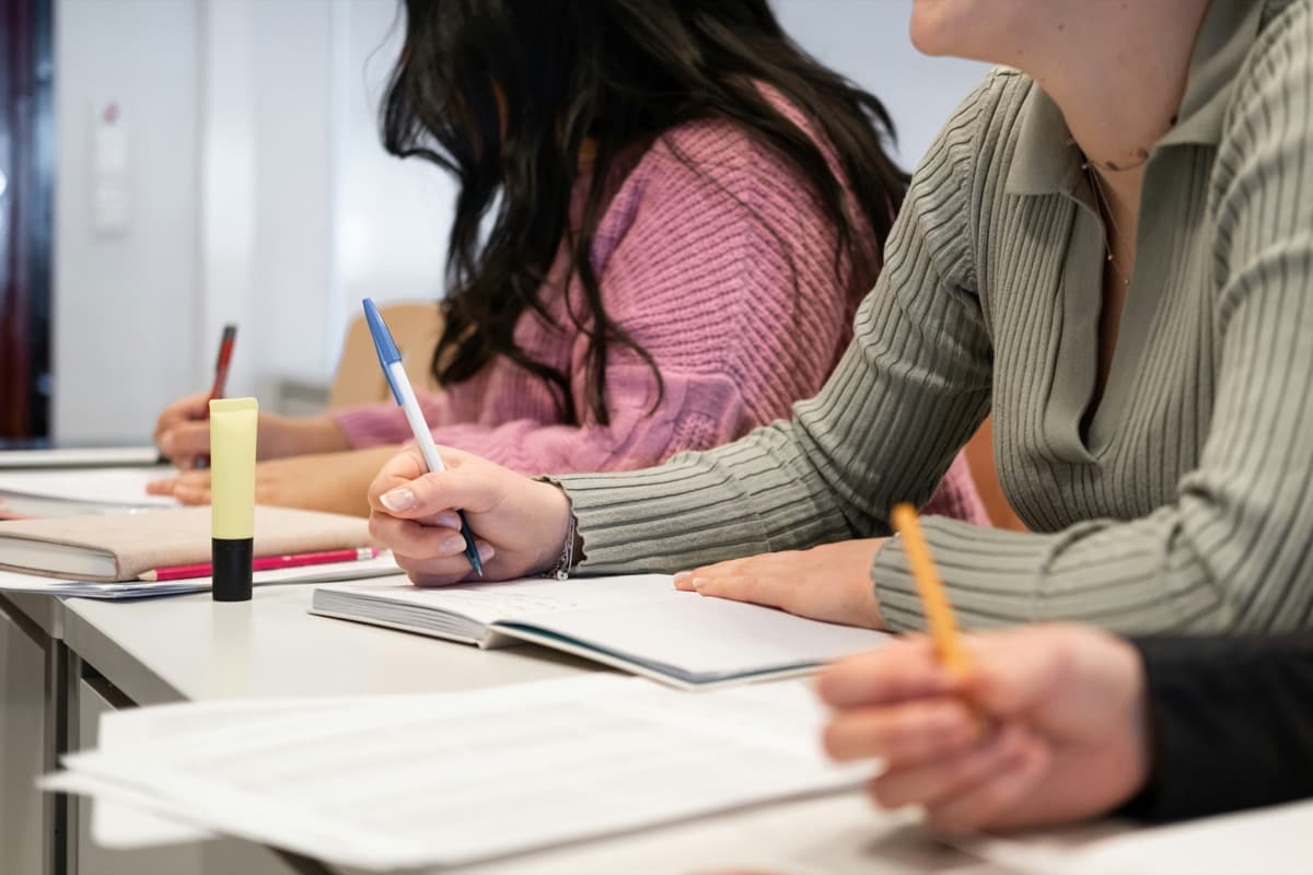 Students seated at a table taking notes in notebooks during a classroom session, emphasizing in-person learning and focused study.