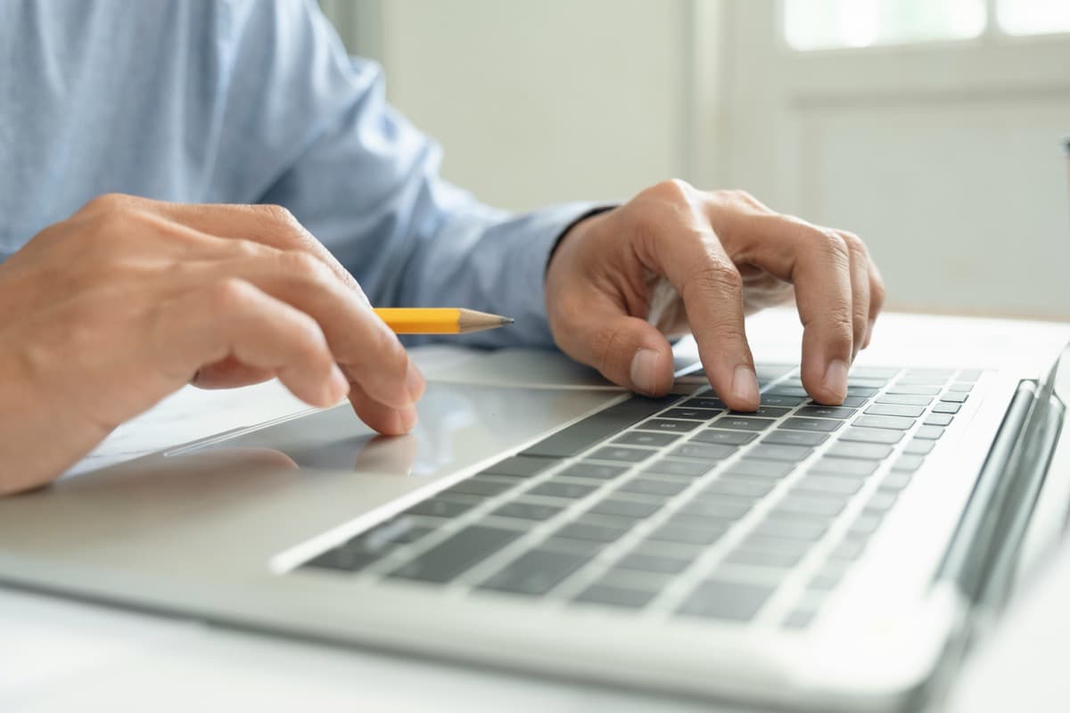 Close-up of a person typing on a laptop with one hand while holding a pencil in the other, suggesting online learning or course registration.
